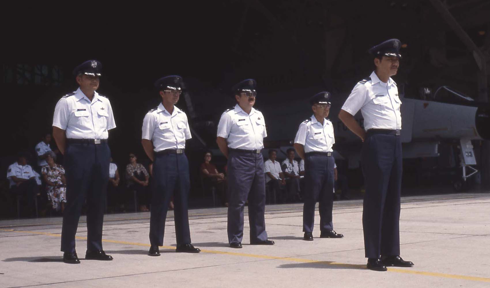 This photograph shows Col John "Saigon" Lee, and his staff, Col Richie "Kuni" Kunichika, Lt Col William "Billy" Mossman, and Lt Col Richard Furuno, in front of a organization formation. It seems to be a retirement or open house ceremony because of the civilians seats in the shade of the hanger (Bldg 3400).